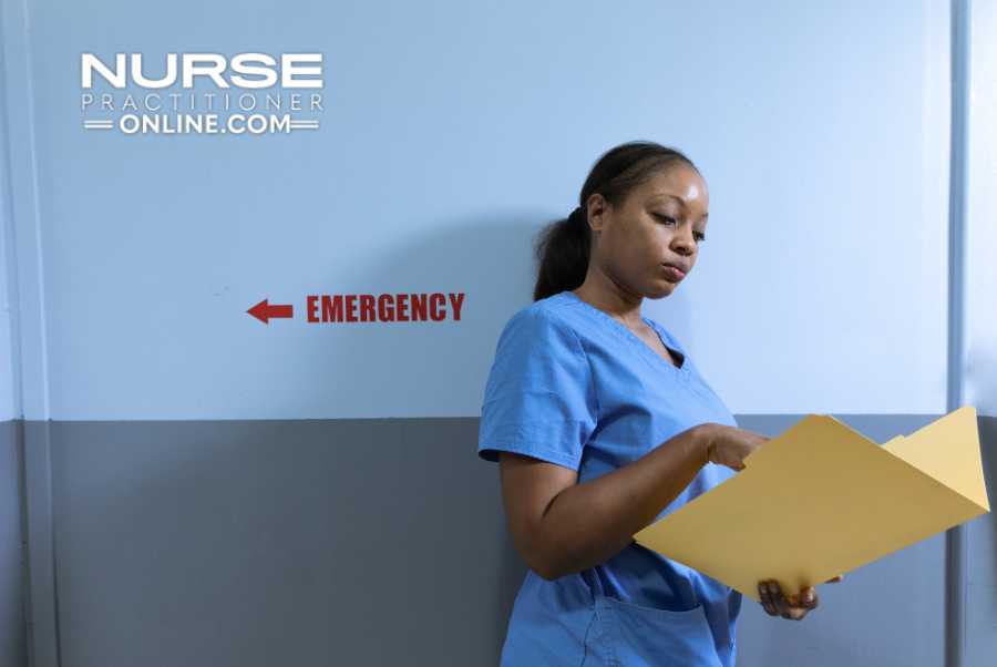 An emergency nurse stands in a hallway and examines a patient's medical file next to the words "Emergency" painted on the wall.